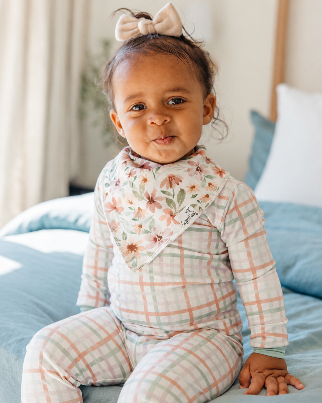 Baby wearing a Copper Pearl Natalie floral bandana bib with a pastel plaid outfit while sitting on a bed.