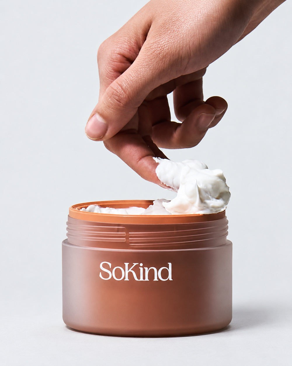 Hand scooping white cream from an open jar of SoKind Love Lines stretch mark balm